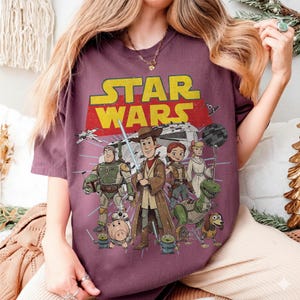 May include: A maroon t-shirt featuring a mashup of Star Wars and Toy Story characters. The design includes the Star Wars logo in yellow and red, with Woody as a Jedi, alongside other characters from both franchises.