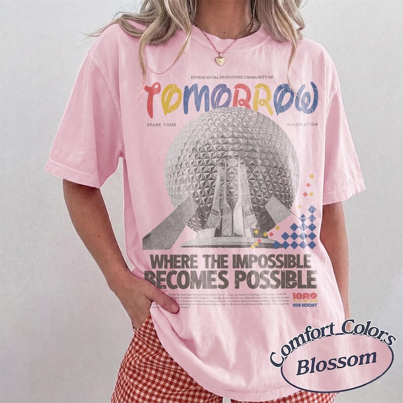 May include: A light pink short-sleeve t-shirt featuring a large graphic of a geodesic dome structure. The text reads "EXPERIMENTAL PROTOTYPE COMMUNITY OF TOMORROW", "SPARK YOUR IMAGINATION", and "WHERE THE IMPOSSIBLE BECOMES POSSIBLE" with colorful accents.