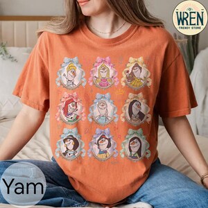 Puede incluir: Camiseta naranja de manga corta con nueve retratos enmarcados de personajes de dibujos animados. Cada retrato est&aacute; en un marco ovalado con un lazo. Los lazos son azules, rosas y amarillos. La camiseta tiene el texto "Yam" en la parte inferior izquierda.