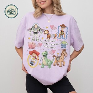 May include: A lilac t-shirt with cartoon characters from a well-known animated film. The design includes Buzz Lightyear, Woody, Jessie, and other characters, with the text "You've Got a Friend in Me."