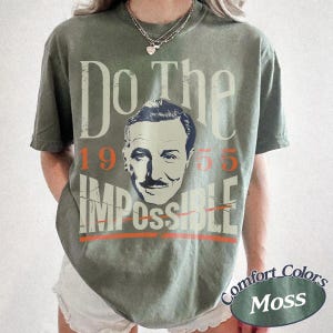 May include: Olive green t-shirt with the text "Do The Impossible" and a portrait of a man. The number "1955" is above the portrait, and the word "Impossible" is below, with a line through it. The shirt has a "Comfort Colors Moss" label.
