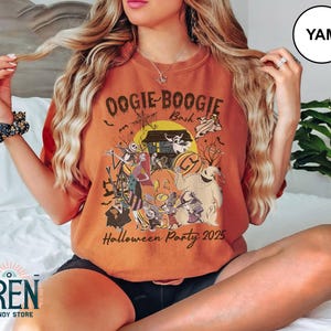 Puede incluir: Camiseta naranja con un dise&ntilde;o tem&aacute;tico de Halloween. El dise&ntilde;o presenta personajes, una casa embrujada, calabazas y el texto "Oogie Boogie Bash Halloween Party 2025". La camiseta tiene un corte holgado.