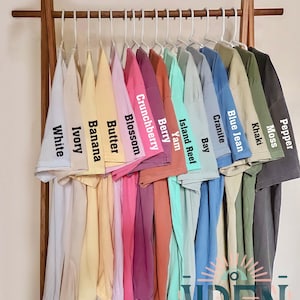K&ouml;nnte beinhalten: Farbkarte mit 12 verschiedenen Farben von Unisex-T-Shirts, darunter Wei&szlig;, Elfenbein, Banane, Butter, Bl&uuml;te, Cranberry, Beere, Yamswurzel, Inselriff, Bucht, Granit, Jeansblau, Khaki und Moos. Die Shirts h&auml;ngen an einem Holzgestell. Der Text "COLOR CHART" und "COMFORT COLORS C1717 UNISEX SHIRT" befindet sich oben im Bild. Der Text "WREN TRENDY STORE" befindet sich unten im Bild.
