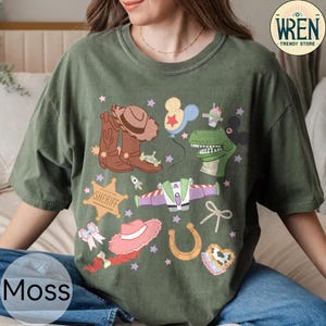 May include: Moss green t-shirt featuring a colorful graphic of cartoon characters and western-themed elements. The design includes a cowboy hat, boots, a sheriff badge, and a dinosaur. The shirt also has the text "WREN TRENDY STORE".