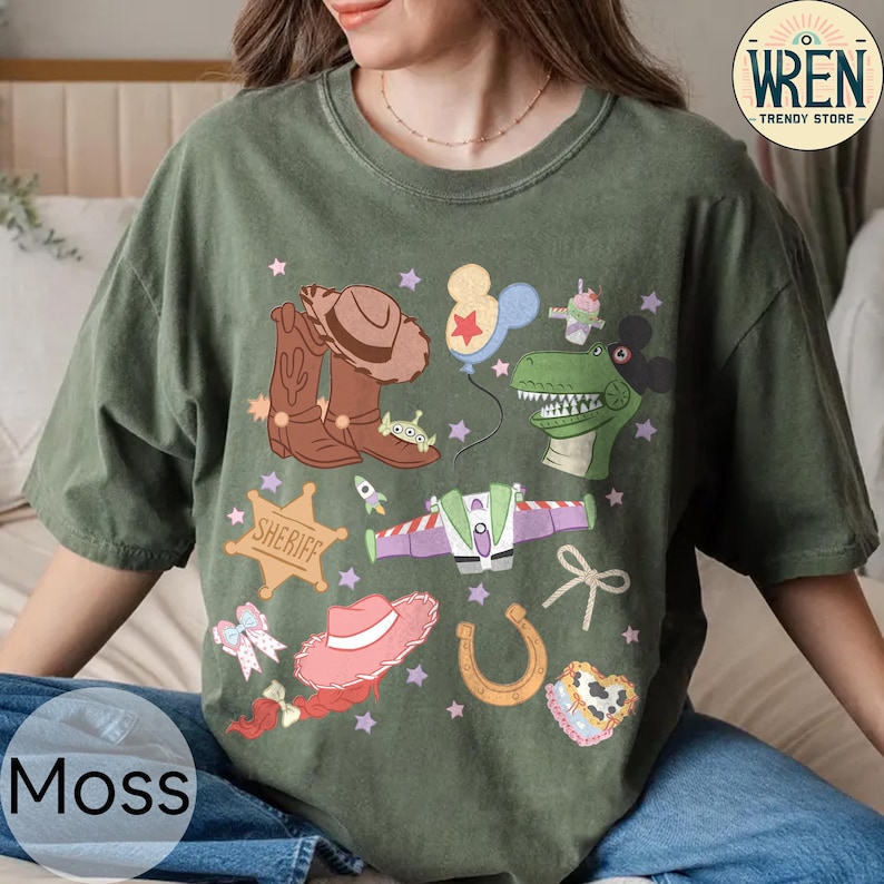 May include: Moss green t-shirt featuring a colorful graphic of cartoon characters and western-themed elements. The design includes a cowboy hat, boots, a sheriff badge, and a dinosaur. The shirt also has the text "WREN TRENDY STORE".