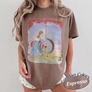 May include: A brown short-sleeve t-shirt featuring a watercolor-style graphic of a person with a red hair bow standing next to a tire swing. The illustration includes a sunset sky, a grassy hill, and a small red heart inside the tire center.