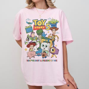 May include: A light pink t-shirt featuring a colorful graphic of characters from the animated film "Toy Story." The design includes Woody, Buzz Lightyear, and other characters, with the text "Toy Story 5" and "You've Got a Friend in Me."