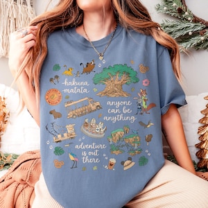 May include: Blue t-shirt with a colorful graphic design featuring cartoon characters, a tree, and the phrases "Hakuna Matata" and "Anyone can be anything." The shirt has a crew neck and short sleeves.