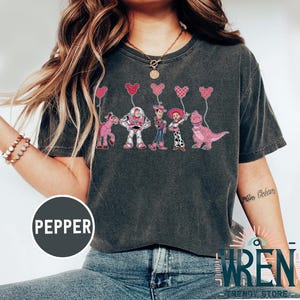 Może przedstawiać: Szary t-shirt z nadrukiem postaci z Toy Story trzymających r&oacute;żowe balony w kształcie serca. Postacie to Woody, Buzz Astral, Jessie i Rex. Tekst "Pepper" jest widoczny w lewym dolnym rogu obrazu.