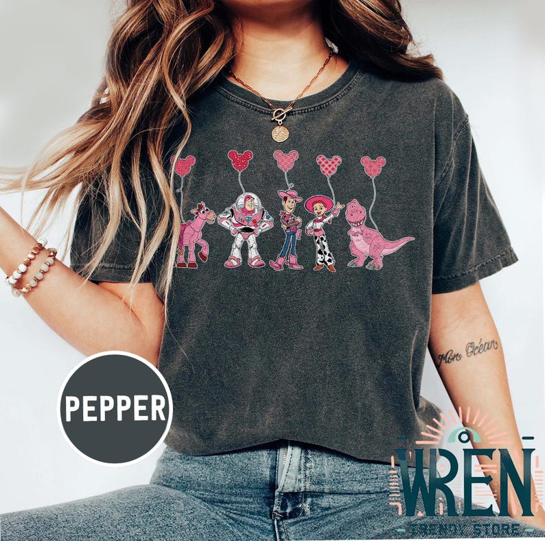 Może przedstawiać: Szary t-shirt z nadrukiem postaci z Toy Story trzymających r&oacute;żowe balony w kształcie serca. Postacie to Woody, Buzz Astral, Jessie i Rex. Tekst "Pepper" jest widoczny w lewym dolnym rogu obrazu.