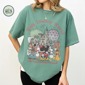 以下が含まれることがあります： ディズニーワールドのグラフィックが特徴の緑色のTシャツ。デザインには、ミッキーマウス、ミニーマウス、ドナルドダック、グーフィー、象徴的なランドマークが含まれています。「ウォルト・ディズニー・ワールド」と「世界で一番幸せな場所」というテキストがあります。