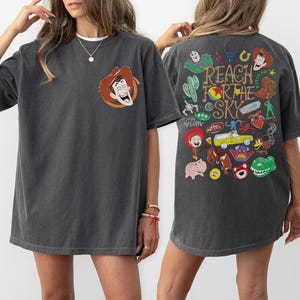 K&ouml;nnte beinhalten: Ein anthrazitfarbenes Kurzarm-T-Shirt mit einer kleinen Grafik von Woody auf der linken Brust. Die R&uuml;ckseite zeigt eine gro&szlig;e, bunte Collage aus Toy Story-Figuren und Symbolen mit dem Text Reach For The Sky in einer stilisierten Schrift.