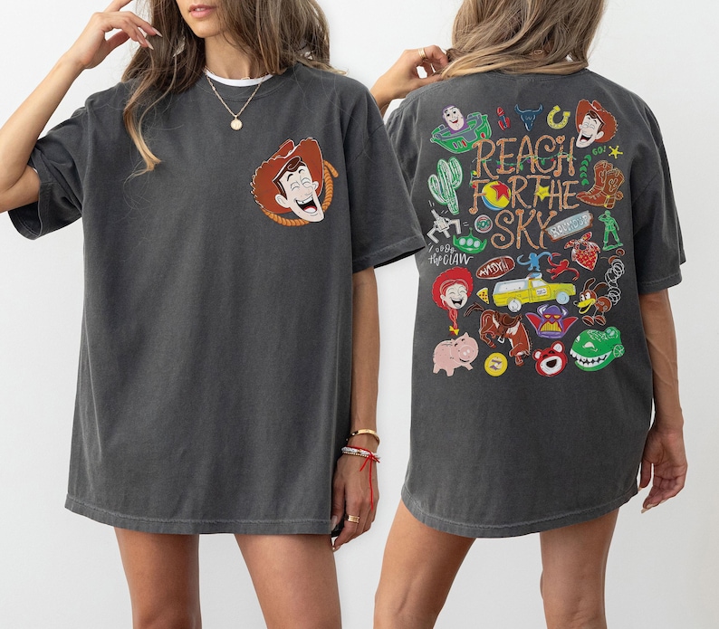 K&ouml;nnte beinhalten: Ein anthrazitfarbenes Kurzarm-T-Shirt mit einer kleinen Grafik von Woody auf der linken Brust. Die R&uuml;ckseite zeigt eine gro&szlig;e, bunte Collage aus Toy Story-Figuren und Symbolen mit dem Text Reach For The Sky in einer stilisierten Schrift.