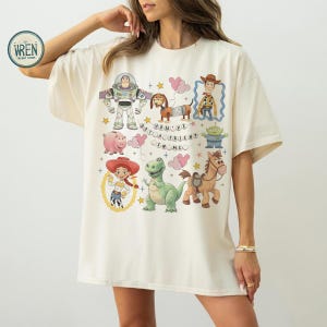 May include: A cream-coloured t-shirt with a colourful Toy Story design. The graphic includes Buzz Lightyear, Woody, Jessie, and other characters, with the phrase "You've Got a Friend in Me".