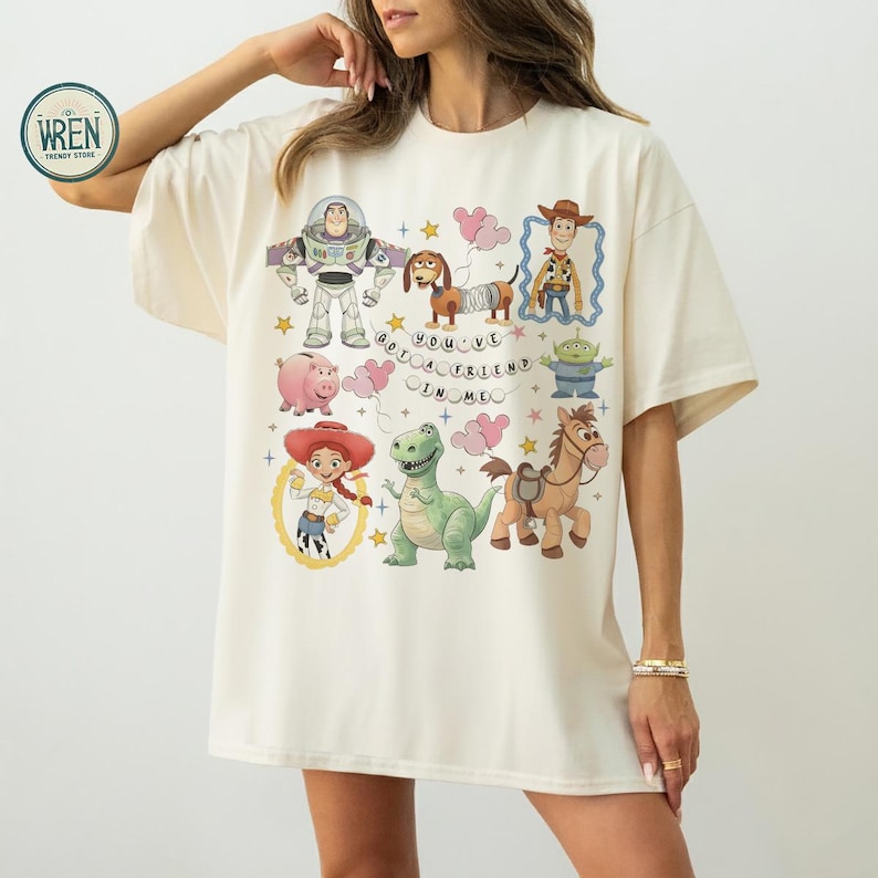 May include: A cream-coloured t-shirt with a colourful Toy Story design. The graphic includes Buzz Lightyear, Woody, Jessie, and other characters, with the phrase "You've Got a Friend in Me".