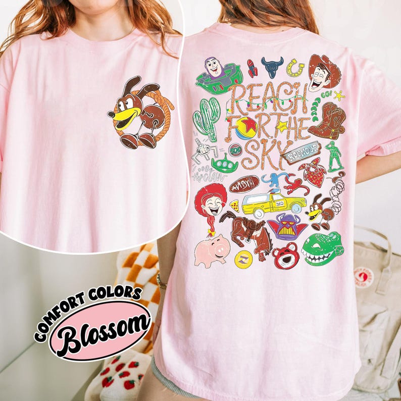 K&ouml;nnte beinhalten: Ein hellrosa T-Shirt mit einer kleinen Grafik eines Hundes mit Lasso auf der linken Brustseite und einer gro&szlig;en, bunten Collage verschiedener Zeichentrickfiguren sowie dem Text "REACH FOR THE SKY" auf der R&uuml;ckseite des Kleidungsst&uuml;cks.
