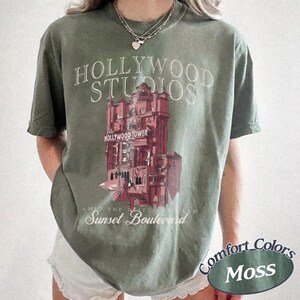 May include: A moss green short-sleeve t-shirt featuring a graphic of a tall building with the text HOLLYWOOD STUDIOS, The Hollywood Tower, and Sunset Boulevard printed in white and red. The shirt has a crew neckline and a relaxed fit.