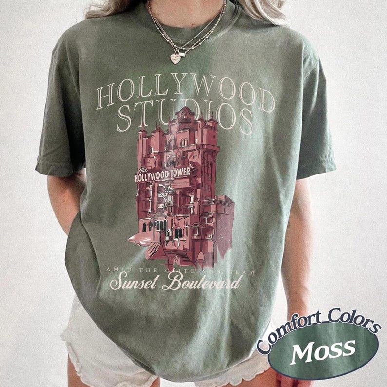 May include: A moss green short-sleeve t-shirt featuring a graphic of a tall building with the text HOLLYWOOD STUDIOS, The Hollywood Tower, and Sunset Boulevard printed in white and red. The shirt has a crew neckline and a relaxed fit.