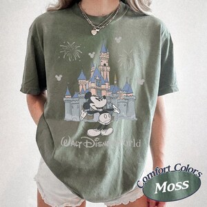 May include: Moss green t-shirt featuring a graphic of Mickey Mouse in front of a castle with fireworks. The text "Walt Disney World" is below the castle. The shirt is a Comfort Colors brand.