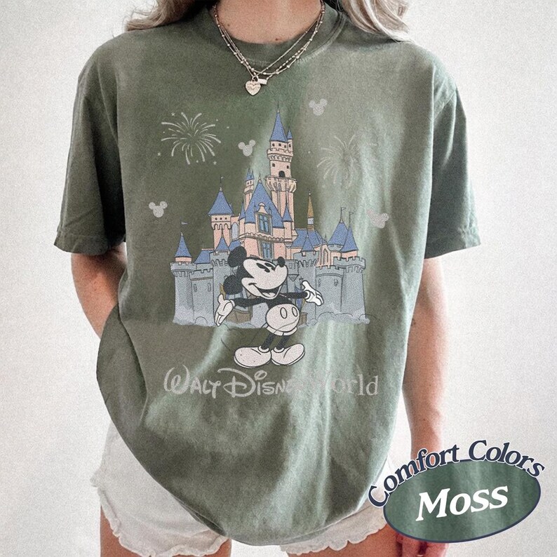 May include: Moss green t-shirt featuring a graphic of Mickey Mouse in front of a castle with fireworks. The text "Walt Disney World" is below the castle. The shirt is a Comfort Colors brand.