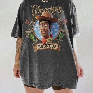 K&ouml;nnte beinhalten: Ein anthrazitfarbenes Kurzarm-T-Shirt mit einem Grafikdruck der Figur Woody aus Toy Story. Das Design zeigt den Schriftzug Woody's Roundup, einen braunen Cowboyhut, einen Sheriffstern, gr&uuml;ne Kakteen und rote Sterne auf blauem Hintergrund.