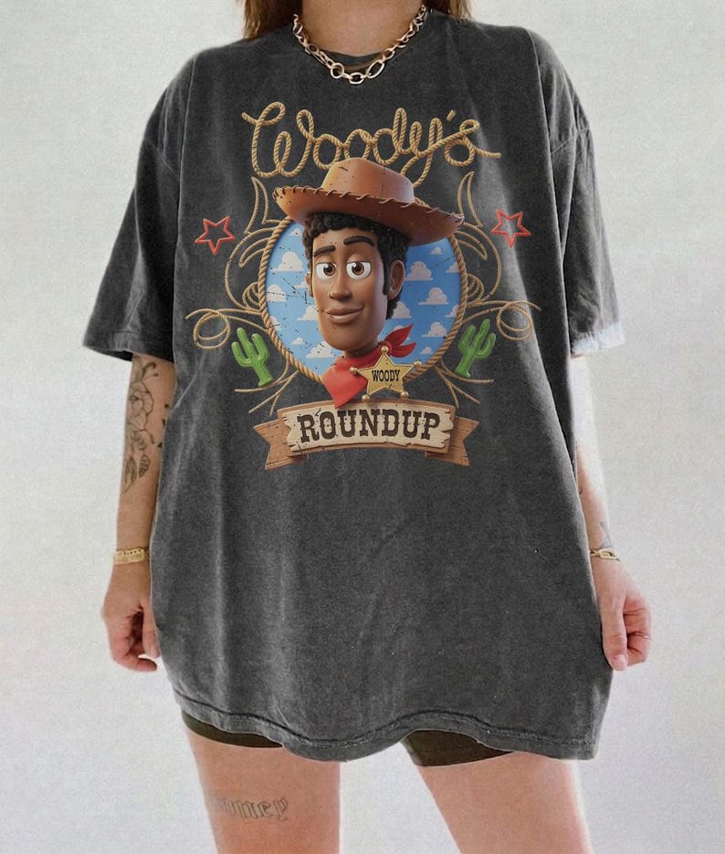 K&ouml;nnte beinhalten: Ein anthrazitfarbenes Kurzarm-T-Shirt mit einem Grafikdruck der Figur Woody aus Toy Story. Das Design zeigt den Schriftzug Woody's Roundup, einen braunen Cowboyhut, einen Sheriffstern, gr&uuml;ne Kakteen und rote Sterne auf blauem Hintergrund.