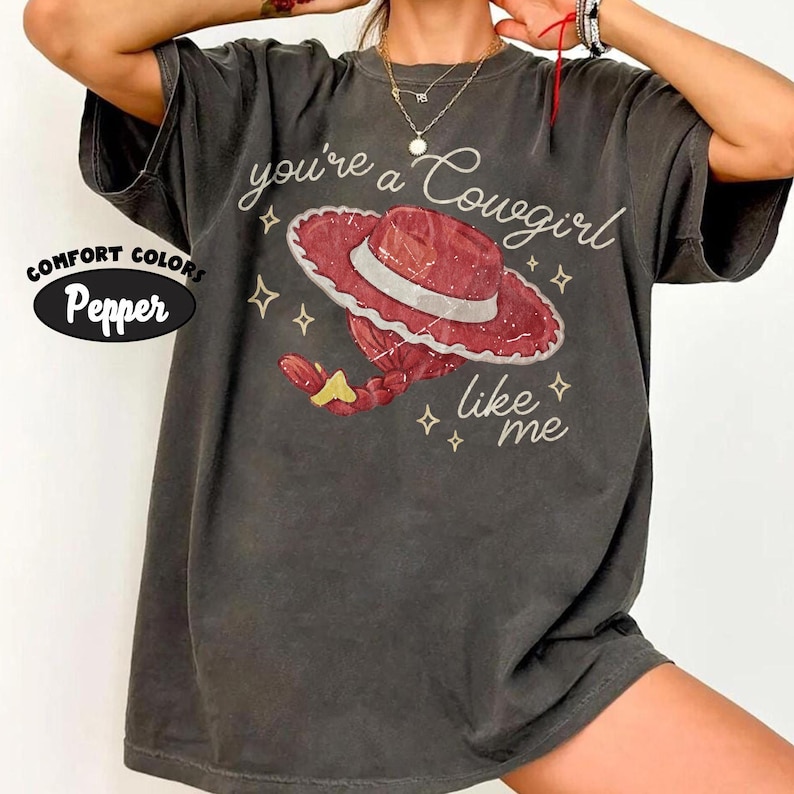 May include: A charcoal gray short-sleeve t-shirt featuring a graphic of a red cowgirl hat with a white band and a red braided ponytail with a yellow hair tie. The text reads you're a Cowgirl like me, surrounded by small white star icons.