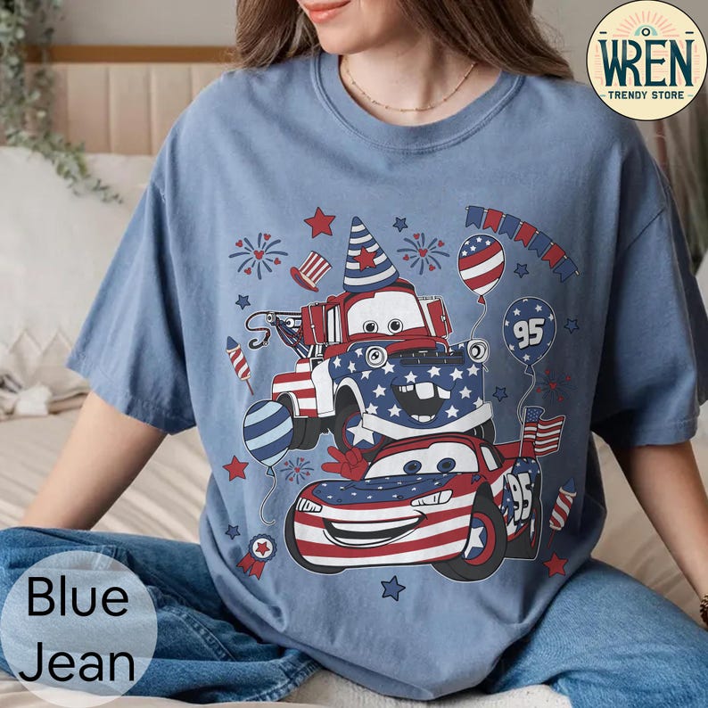 Puede incluir: Camiseta azul vaquero con un dise&ntilde;o patri&oacute;tico con coches de dibujos animados, estrellas y fuegos artificiales. El dise&ntilde;o incorpora los colores rojo, blanco y azul, con el n&uacute;mero "95" en un globo y un coche. La camiseta tiene un corte holgado.