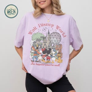以下が含まれることがあります： ウォルト・ディズニー・ワールドのグラフィックがプリントされた薄紫色のTシャツ。「ウォルト・ディズニー・ワールド」と「The happiest place on earth」の文字と、ディズニーのキャラクターやランドマークのイラストが描かれています。