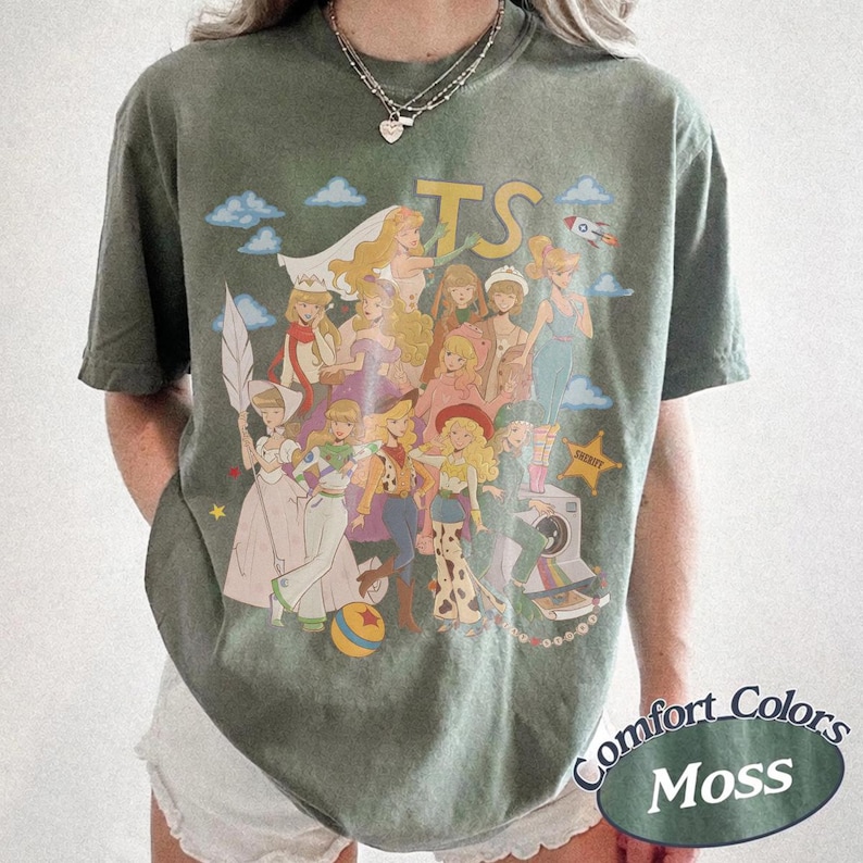 May include: A moss green short-sleeve t-shirt featuring a large graphic print of various animated characters in diverse outfits. The design includes the letters TS at the top, a small rocket, clouds, and a yellow star with the word Sheriff printed on it.