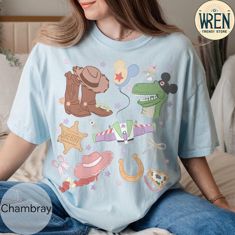 May include: Light blue t-shirt with a colorful graphic featuring cowboy boots, a sheriff's badge, a pink hat, a horseshoe, and a dinosaur. The shirt also has the word "Chambray" printed on it.
