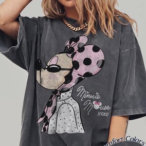 May include: Dark gray t-shirt featuring a Minnie Mouse graphic with a pink polka dot bow and scarf. The text "Minnie Mouse xoxo" is printed on the shirt. The shirt is labeled "Comfort Colors Pepper".