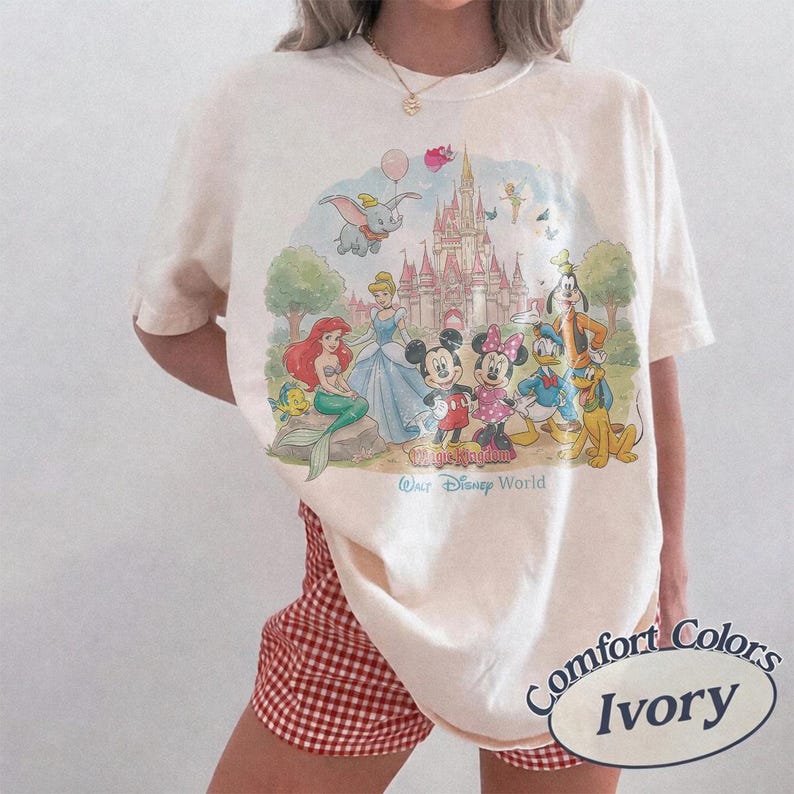 May include: An ivory short-sleeve t-shirt featuring a colorful graphic print of Disney characters including Mickey Mouse, Minnie Mouse, Ariel, Cinderella, Dumbo, and Goofy in front of a castle with the text Walt Disney World printed below.