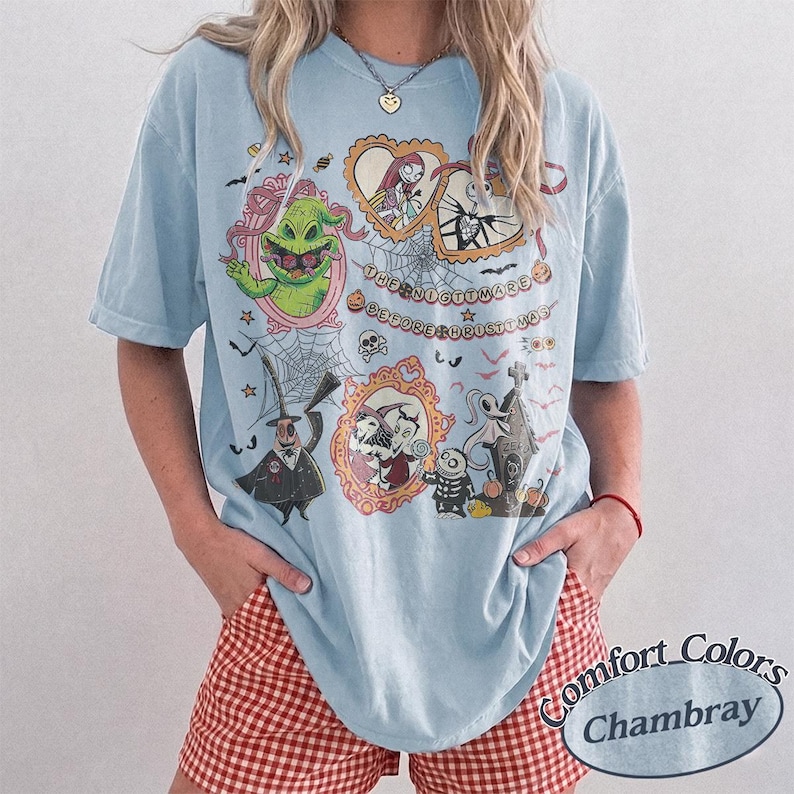 May include: Light blue t-shirt featuring characters from "The Nightmare Before Christmas." The graphic includes images of Jack Skellington, Sally, and Oogie Boogie, with the text "The Nightmare Before Christmas." The shirt is a Comfort Colors Chambray.
