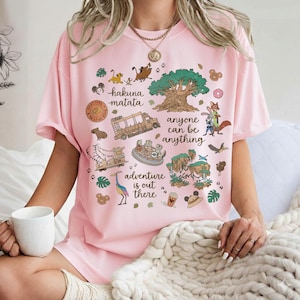 May include: Pink t-shirt featuring a colorful graphic with the text "Hakuna Matata", "Anyone Can Be Anything", and "Adventure is Out There". The design includes cartoon characters, a tree, and other themed elements.