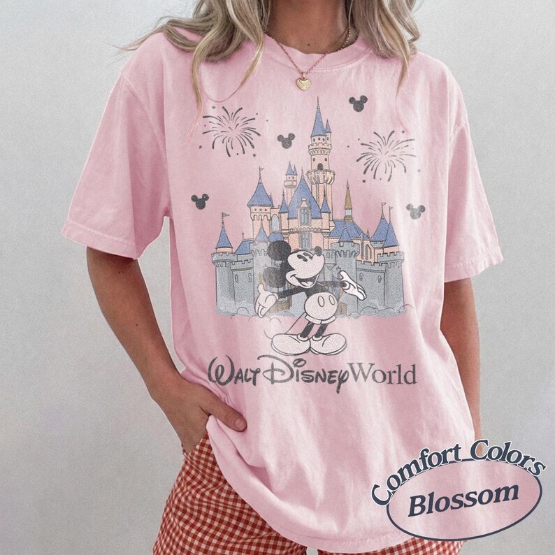 May include: Pink t-shirt featuring a graphic of Mickey Mouse in front of Cinderella's castle at Walt Disney World. The shirt has the text "Walt Disney World" and "Comfort Colors Blossom".