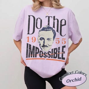 May include: Orchid-colored t-shirt with the text "Do The Impossible 1955" and a portrait. The shirt is a Comfort Colors brand. The design features a vintage aesthetic.