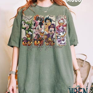May include: Moss green t-shirt featuring a Halloween design with cartoon characters. The design includes Goofy, Minnie Mouse, Mickey Mouse, and Donald Duck, each in costume, with pumpkins and the text "Spooky Season".