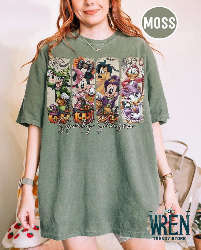 May include: Moss green t-shirt featuring a Halloween design with cartoon characters. The design includes Goofy, Minnie Mouse, Mickey Mouse, and Donald Duck, each in costume, with pumpkins and the text "Spooky Season".
