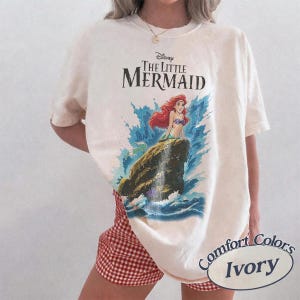 May include: An ivory-colored short-sleeve t-shirt featuring a graphic print of Ariel from Disney's The Little Mermaid sitting on a rock amidst blue ocean waves. The text Disney The Little Mermaid is printed in black above the character illustration.