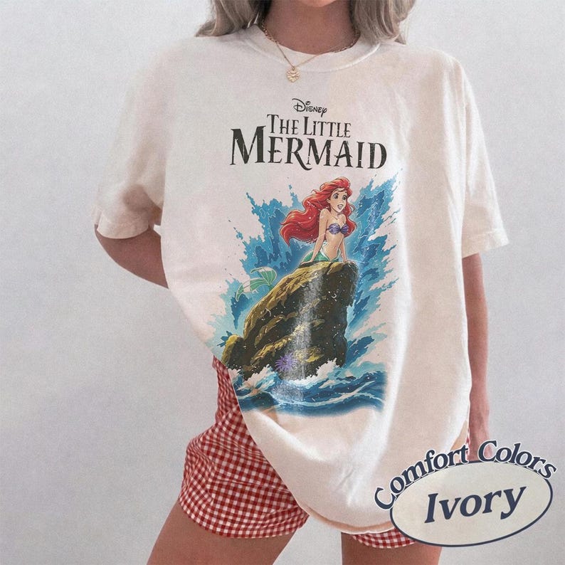 May include: An ivory-colored short-sleeve t-shirt featuring a graphic print of Ariel from Disney's The Little Mermaid sitting on a rock amidst blue ocean waves. The text Disney The Little Mermaid is printed in black above the character illustration.