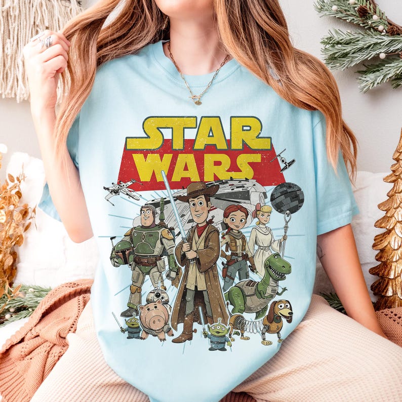 May include: Light blue t-shirt featuring a graphic of Star Wars characters, including Woody, Buzz Lightyear, and others. The Star Wars logo is in yellow and red. The shirt is a comfortable, casual style.