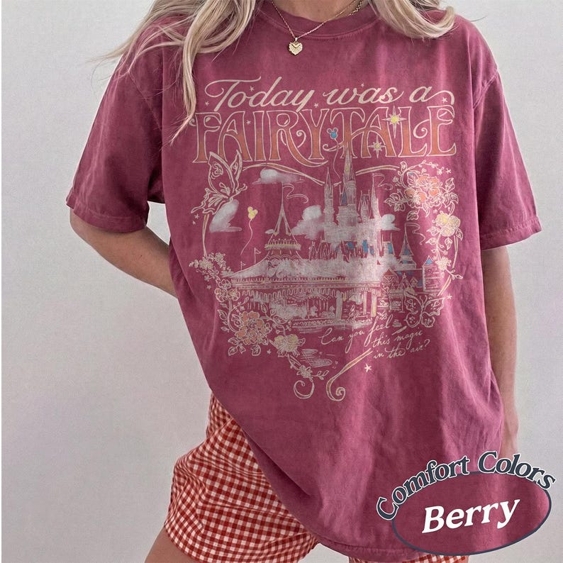 Puede incluir: Una camiseta de manga corta color baya con un estampado gr&aacute;fico de un castillo, una mariposa y elementos florales. El texto dice Today was a FAIRY TALE y Can you feel this magic in the air? La camiseta tiene cuello redondo y un corte holgado.