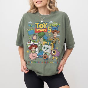 May include: Olive green t-shirt featuring a colorful graphic of characters from the animated film "Toy Story 5". The design includes Woody, Buzz Lightyear, and other characters. The text "TOY STORY 5" is displayed prominently, with the phrase "YOU'VE GOT A FRIEND IN ME" below.