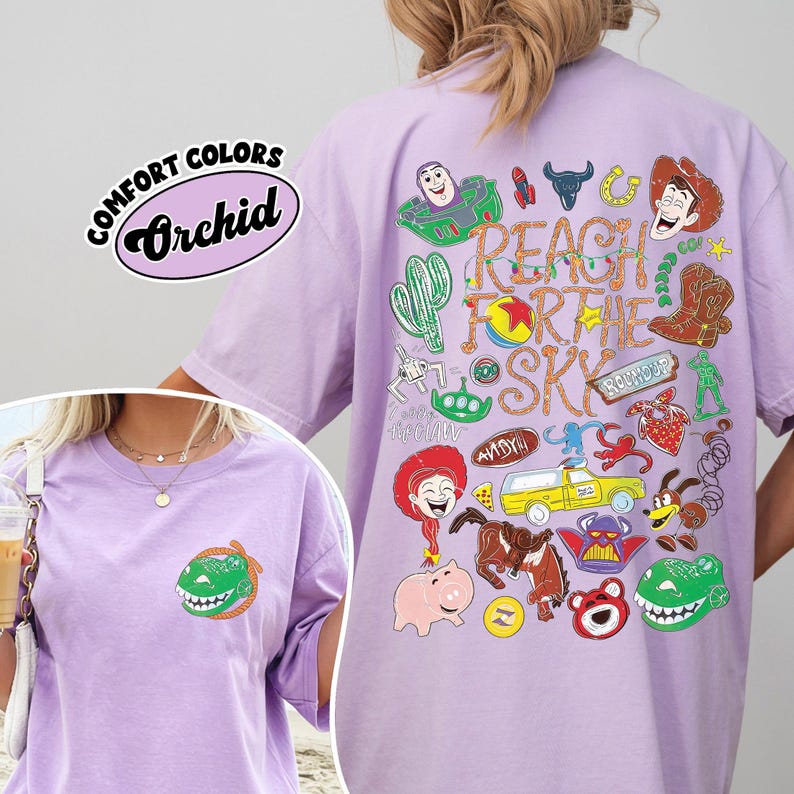 K&ouml;nnte beinhalten: Ein hellviolettes T-Shirt mit einer kleinen gr&uuml;nen Dinosaurier-Grafik auf der Brust. Die R&uuml;ckseite zeigt eine gro&szlig;e, bunte Collage aus verschiedenen Zeichentrickfiguren und den Schriftzug Reach For The Sky in einer dekorativen Schrift.