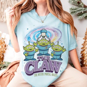 以下が含まれることがあります： 水色のTシャツ。3つ目の緑色の宇宙人が青い服を着て、クレーンゲームの下にいます。デザインには銀河と、紫色の文字で「THE CLAW」と「THE MYSTIC PORTAL AWAITS」が含まれています。