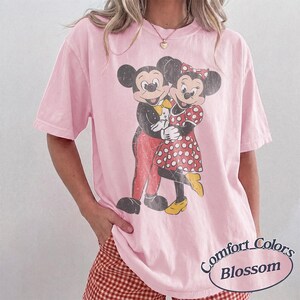 May include: A light pink short-sleeve t-shirt featuring a distressed graphic of Mickey and Minnie Mouse embracing. Mickey wears red pants and a yellow bow tie, while Minnie wears a red and white polka dot dress and yellow shoes. Text reads Comfort Colors Blossom.