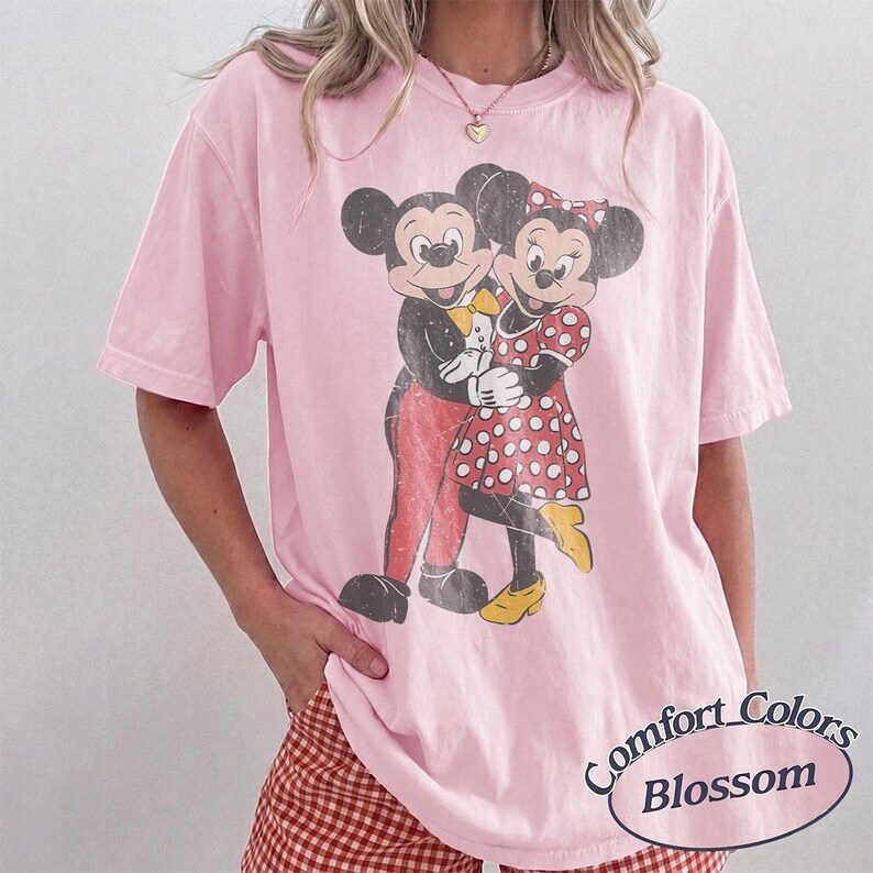 May include: A light pink short-sleeve t-shirt featuring a distressed graphic of Mickey and Minnie Mouse embracing. Mickey wears red pants and a yellow bow tie, while Minnie wears a red and white polka dot dress and yellow shoes. Text reads Comfort Colors Blossom.