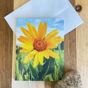 May include: A greeting card featuring a vibrant yellow sunflower painting against a blue sky, with a white envelope. The artwork showcases the flower's detailed petals and center, set against a green field. A small, textured stone rests nearby.