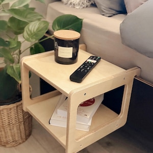 Puede incluir: Una mesita de noche de madera clara con dos estantes. Una vela negra con tapa de corcho, un mando a distancia y dos libros están sobre la mesa. Una planta verde y una cama están en el fondo.